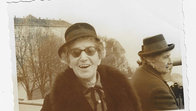 Foto von lachender Person mit Mantel, Hut und Brille, im Hintergrund eine weitere Person, Bäume und ein Haus.