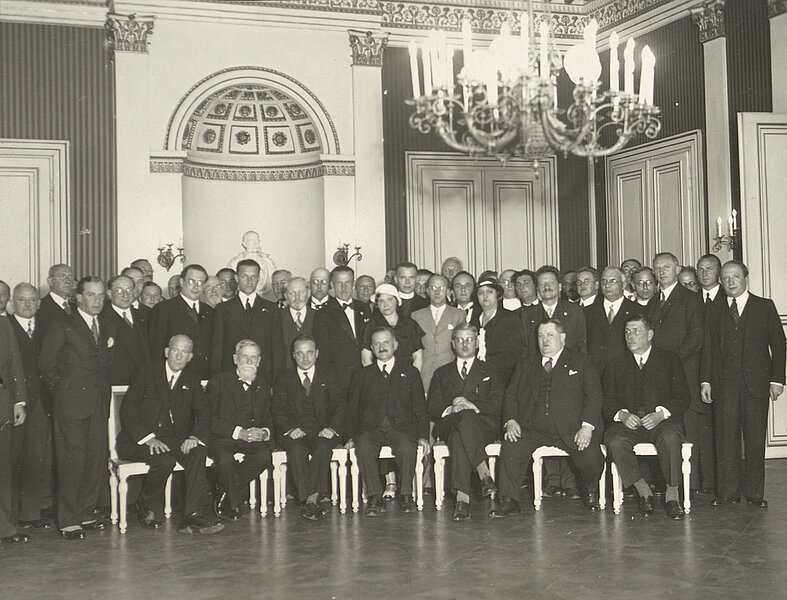 Eine große Gruppe posiert in edlem Saal für ein Foto, schwarz-weiß