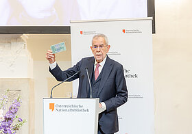 Person in dunklem Anzug hält Rede an Pult, daneben große Blumendekoration, hälten Geldschein in die Luft..