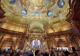 Vielen Personen bei einem Champagner-Empfang in einem großen barocken Saal. 