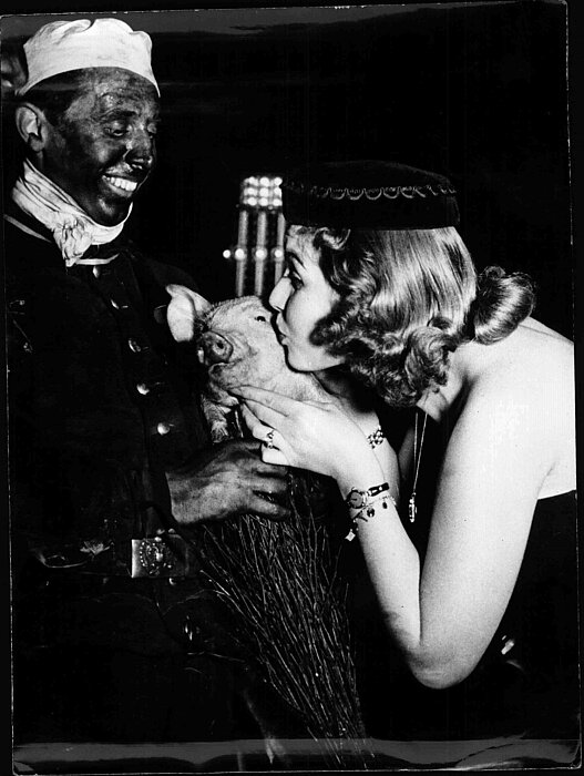 Schwarz-weißes Foto von einem Rauchfangkehrer, der ein Schwein hält. Eine Person küsst das Schwein auf die Wange.