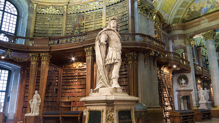 Foto von Statue in barockem Saal mit hohen Bücherregalen und Fresken.