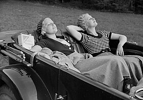 Schwarz-weißes Foto von zwei Personen, die sich in einem offenen Cabrio sonnen.