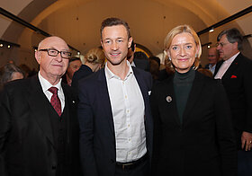 Drei Personen in eleganter Kleidung stehen nebeneinander.