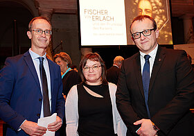 Drei bebrillte Personen in eleganter Kleidung. Im Hintergrund Leinwand mit Projektion. 