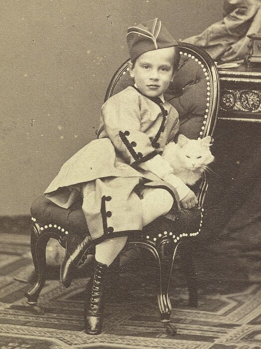 Schwarz-weißes Foto von kleinem Kind in edler Kleidung auf Sessel mit weißer Katze.