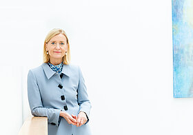 Porträtfoto von einer blonden Frau in hellblauer Weste neben einem blauen Gemälde