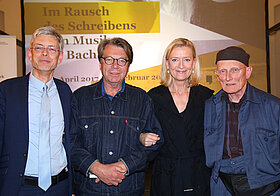 Vier Personen in eleganter Kleidung lächelnd nebeneinander. Im Hintergrund eine Leinwand.