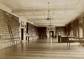 Sepia-Foto eines großen Saals mit Bücherregalen an den Wänden und Tischvitrinen.