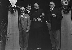 Fritz Grünbaum und Egon Fridell stehen mit Männern die eine Büste halten auf der bühne