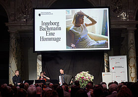 Musik-Trio auf einer Bühne vor Publikum, dahinter Leinwand mit Foto von Ingeborg Bachmann..
