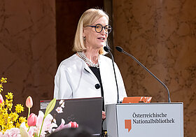 Person in eleganter Kleidung spricht an einem Pult.