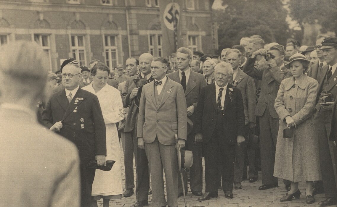 Schwarz-weißes Foto von Menschenmenge in Anzügen oder Mänteln, im Hintergrund eine Hakenkreuzflagge.