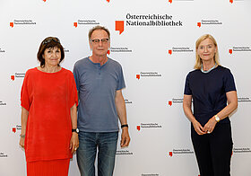 Drei Personen stehen lächelnd vor einer Logowand.
