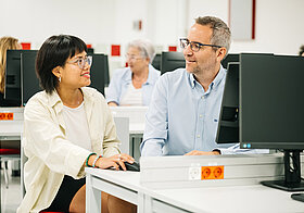 Eine jüngere Person und eine älterer Person sitzen an einem Computer.