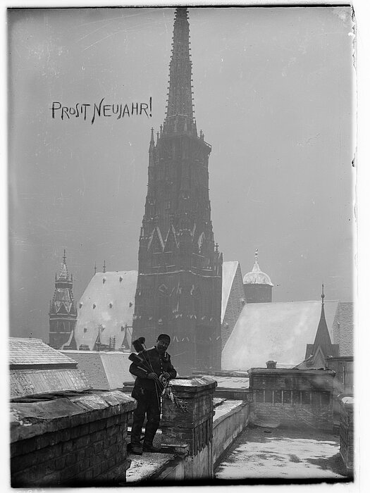 Schwarz-weißes Foto von Rauchfangkehrer auf einem Dach, im Hintergrund eine schneebedeckte Kirche. Im "Himmel" steht "Prosit Neujahr!".