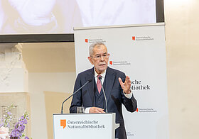 Person in dunklem Anzug hält Rede an Pult, daneben große Blumendekoration.