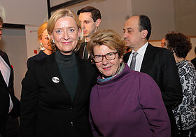 Eine blonde Person und eine Person mit Brille lächeln in die Kamera.