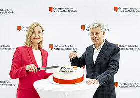 Zwei Personen in Anzügen stehen vor einer Wand mit dem Logo der Österreichischen Nationalbibliothek, zwischen ihnen steht eine Torte mit der Aufschrift "Zehn Jahre Literaturmuseum", die angeschnitten wird.