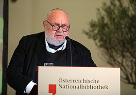 Bebrillte Person in dunkler Kleidung am Rednerpult.