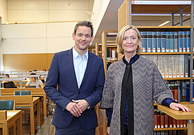 Zwei Personen posieren in Saal mit Bücherregalen und Schreibtischen.