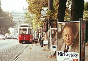 Foto einer Straßenbahnhaltestelle mit Bäumen, Menschen und Plakaten auf denen ein Mann abgebildet ist.
