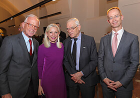 Drei Personen in Anzügen und eine Person in lilafarbenem Kleid.