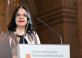 Person in brauner Jacke spricht an einem Rednerpult.