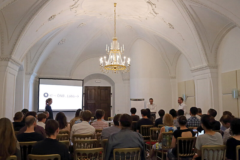 Menschen sitzen vor Leinwand in Raum mit Kronleuchter, ÖNB Labs Symposium, Begrüßung durch Max Kaiser