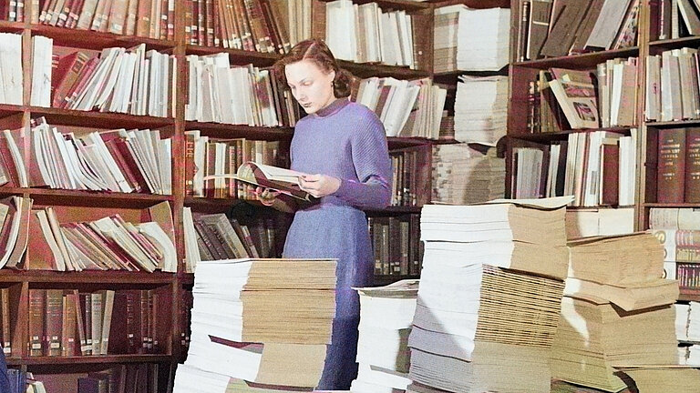 Mensch steht vor einem Bücherregal und liest ein Buch. 