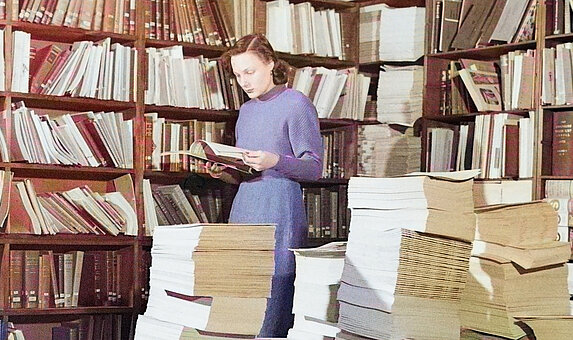 Mensch steht vor einem Bücherregal und liest ein Buch. 