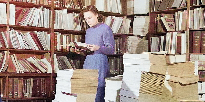 Mensch steht vor einem Bücherregal und liest ein Buch. 