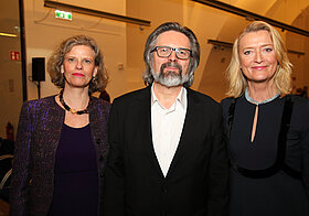 Zwei Personen in eleganter Kleidung und bebrillte Person im Anzug posieren lächelnd.