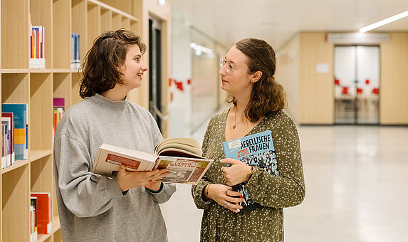 Foto von zwei Personen, die sich unterhalten und dabei Bücher halten.