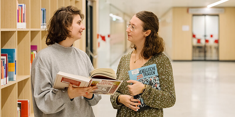 Foto von zwei Personen, die sich unterhalten und dabei Bücher halten.