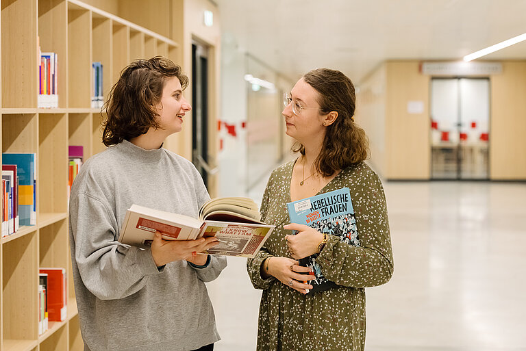 Foto von zwei Personen, die sich unterhalten und dabei Bücher halten.