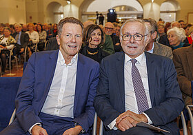 Zwei Personen sitzen in Publikum und schauen in die Kamera.
