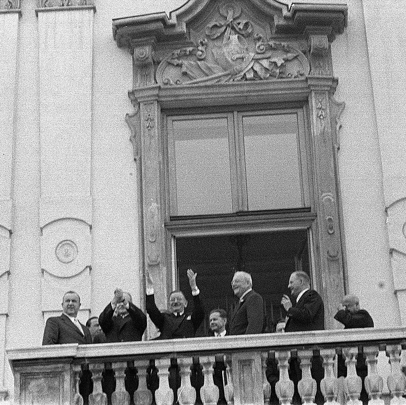 Männer in Anzügen stehen erfreut auf einem Balkon, zwei heben die Arme, schwarz-weiß
