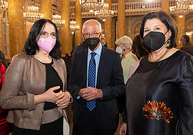Drei Personen in eleganter Kleidung stehen im Marmorsaal. Im Hintergrund prachtvolle Kronleuchter.