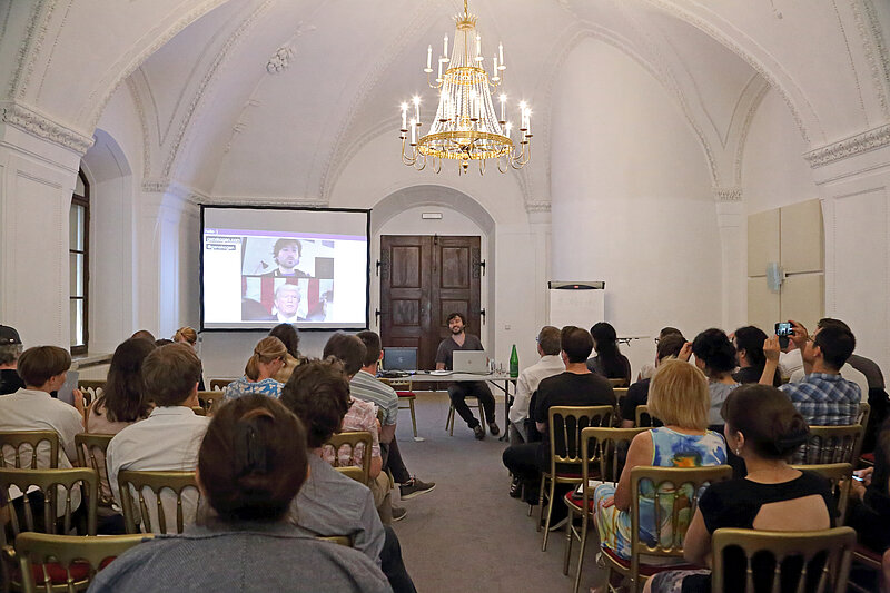 Menschen sitzen vor Leinwand in Raum mit Kronleuchter, ÖNB Labs Symposium, Keynote von Gene Kogan