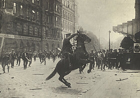 Schwarz-weißes Foto von Polizist auf einem sich aufbäumenden Pferd, einen Stock erhoben. Im Hintergrund Stadt mit Menschenmenge.