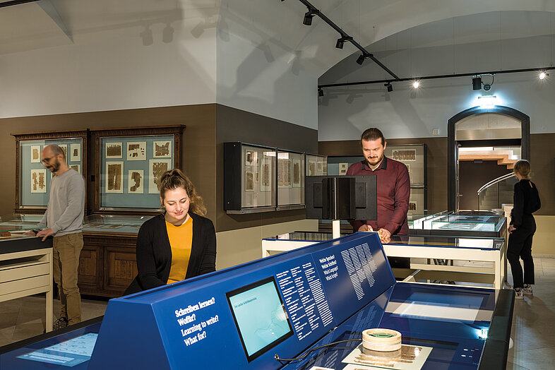 Museumsraum mit Besuchenden, in der Mitte sind Tische mit Bildschirmen zu sehen.