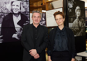 Zwei Personen im Anzug posieren vor Museumsregalen mit schwarz-weißen Fotos.