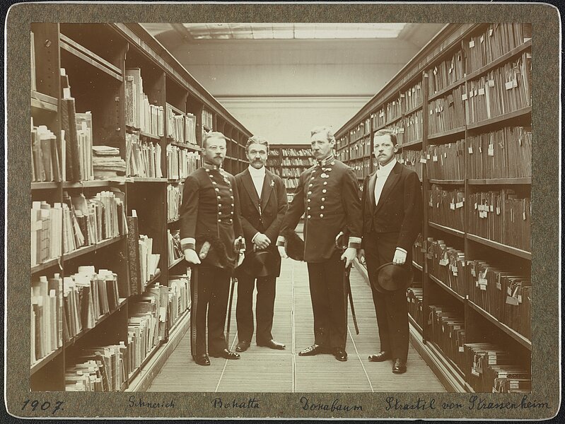 Vier Männer in Uniform bzw. Frack stehen zwischen Bücherregalen, sepia