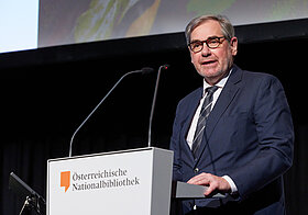 Bebrillte Person im Anzug am Rednerpult.