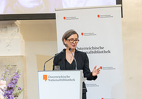 Person in dunklem Anzug hält Rede an Pult, daneben große Blumendekoration.