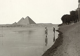 Schwarz-weißes Foto von zwei Personen die am Ufer im Wasser stehen, im Hintergrund Pyramiden