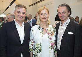 Zwei Person im Anzug und eine Person mit Blumenblazer posieren lächelnd.