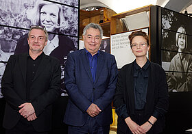 Drei Personen posieren vor Museumsregalen mit schwarz-weißen Fotos