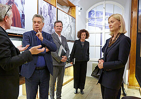 Eine Gruppe von Menschen unterhalten sich zwischen Museumsregalen. Im Hintergrund Foto von Ingeborg Bachmann. 